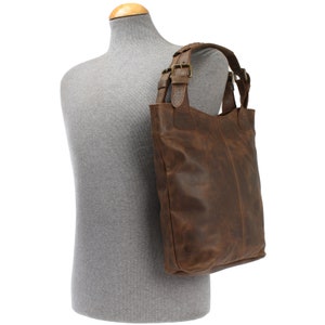 May include: A brown leather tote bag with double buckles on the straps. The bag has a distressed look and is worn on a gray mannequin.