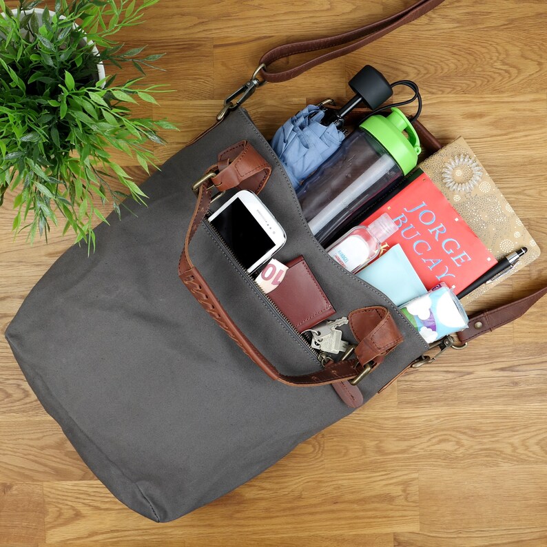 May include: A gray canvas tote bag with brown leather straps and a zippered pocket. The bag is filled with various items, including a phone, a wallet, a book, a water bottle, and a hand sanitizer.