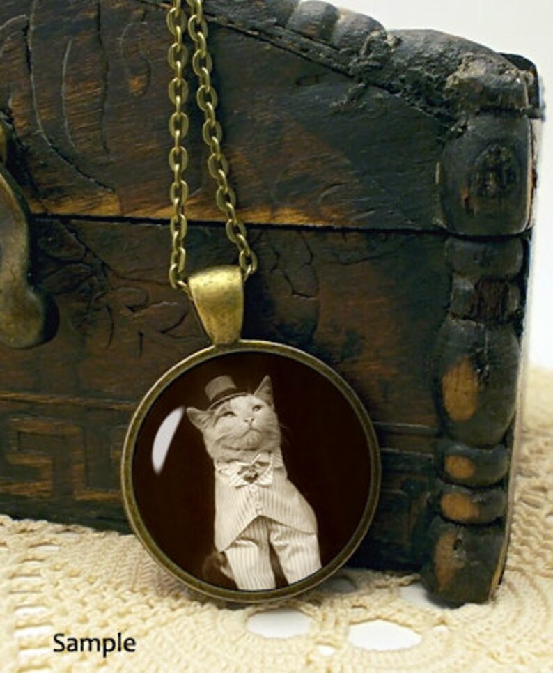 K&ouml;nnte beinhalten: Eine bronzene Halskette mit einem runden Glasanh&auml;nger, der ein Schwarzwei&szlig;foto einer Katze mit Zylinder und Fliege zeigt.