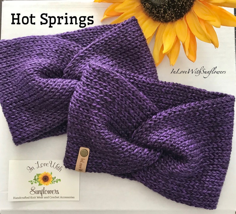 May include: Purple knitted headband with a twisted knot design. The headband is handmade and has a small brown leather tag with the text "In Love With" on it.