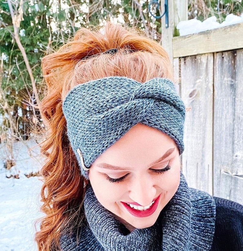 May include: A gray and blue knit headband with a twisted knot design. The headband is worn by a person with red hair.