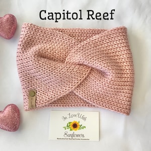 May include: A pink knitted headband with a knotted design. The headband has a brown leather tag with the text "In Love With Sunflowers" on it. The text "Capitol Reef" is written in black above the headband.
