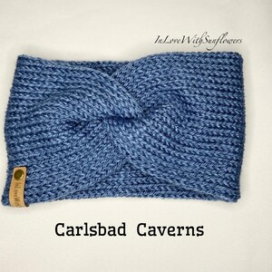 May include: A blue knitted headband with a knotted design. The headband has a brown leather tag with the text "In Love With" on it. The text "Carlsbad Caverns" is written below the headband.