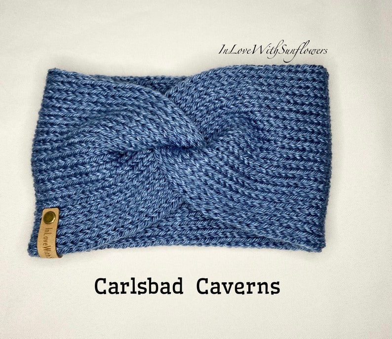 May include: A blue knitted headband with a knotted design. The headband has a brown leather tag with the text "In Love With" on it. The text "Carlsbad Caverns" is written below the headband.