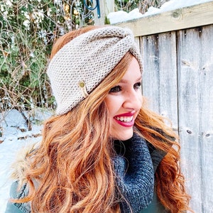 May include: A beige knitted headband with a knotted detail and a small gold button. The headband is worn by a person with long, wavy, red hair.