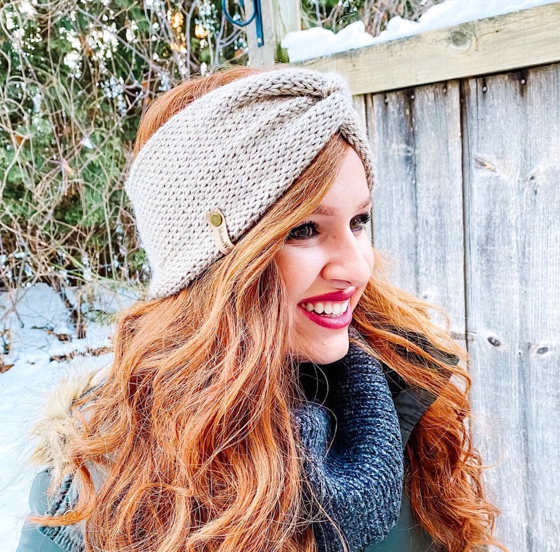 May include: A beige knitted headband with a knotted detail and a small gold button. The headband is worn by a person with long, wavy, red hair.