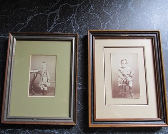 May include: Two framed sepia-toned photographs of young boys. The boy in the left photo is standing next to a table and holding a small object. The boy in the right photo is sitting on a stool. Both photos have a green mat and a wooden frame.
