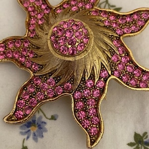 May include: A gold-toned starburst brooch with pink rhinestones. The brooch features a central circular design and radiating points, all encrusted with sparkling pink stones. The brooch is a vintage accessory.