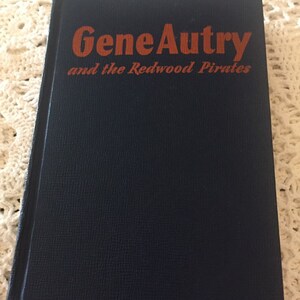 May include: A dark blue book with the title "Gene Autry and the Redwood Pirates" in orange lettering.