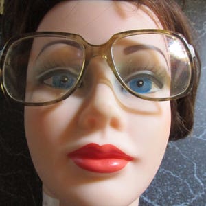 May include: A close-up of a woman's face wearing brown-rimmed glasses with a red lipstick. The glasses have a retro style and the woman has a neutral expression.