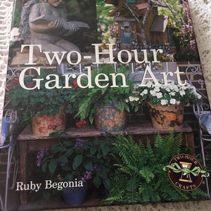 May include: A book titled "Two-Hour Garden Art" by Ruby Begonia, featuring a whimsical garden scene with a birdhouse, potted plants, and a cherub statue reading a book.