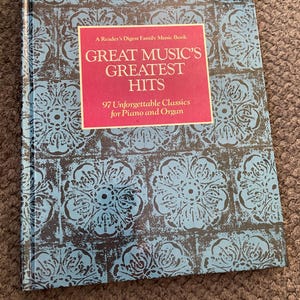 May include: A Reader's Digest Family Music Book titled "Great Music's Greatest Hits." The book cover features a blue and black floral pattern. A pink rectangle on the cover displays the title and the text "97 Unforgettable Classics for Piano and Organ."