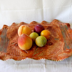 Op de afbeelding: Een rustieke, bruine keramische serveerschaal met een gegolfde rand, met een getextureerd ontwerp. De schaal bevat een verscheidenheid aan fruit, waaronder perziken, pruimen en groene pruimen.