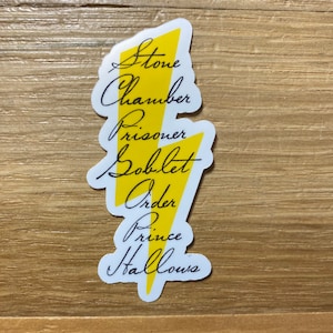 May include: A white sticker in the shape of a yellow lightning bolt with black text. The text includes the words "Stone", "Chamber", "Prisoner", "Goblet", "Order", "Prince", and "Hallows". The sticker is on a light brown wooden surface.