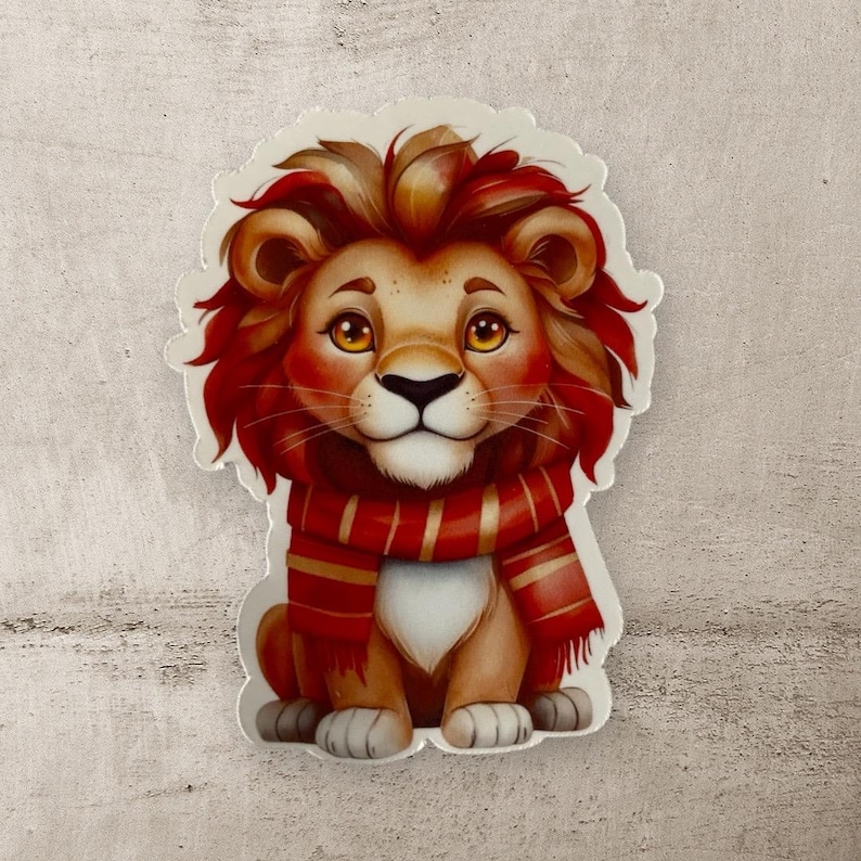 May include: A cartoon lion cub wearing a red and gold striped scarf. The lion has a fluffy mane and is sitting with its paws crossed.