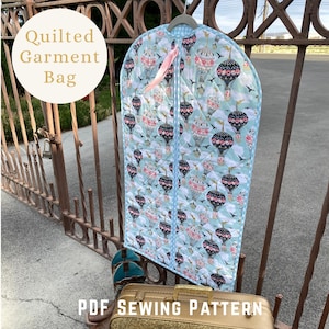 May include: A quilted garment bag featuring a light blue background with a repeating pattern of hot air balloons, clouds, and small birds. The bag has a central zipper with a pink ribbon pull and light blue and white checkered trim edges.