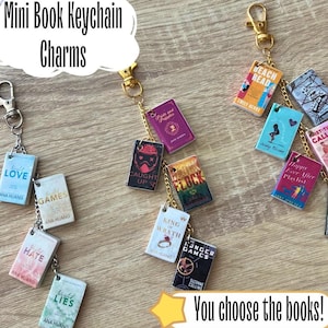May include: Miniature book charms on keychains. The charms feature book covers with titles like "Love," "Games," and "The Hunger Games." The keychains have gold-coloured clasps and chains. The text "You choose the books!" is also visible.