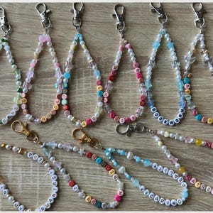 Pode incluir: Uma coleção de chaveiros de contas com várias cores e designs. Alguns chaveiros apresentam palavras soletradas com contas de letras, incluindo "READING", "BOOKWORM" e "ESCAPE". Os chaveiros têm fechos prateados e dourados.