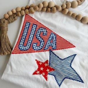 May include: A white t-shirt featuring a red stitched triangular pennant with the blue and white gingham letters USA, accompanied by a red star with white stars and a larger blue star with horizontal blue stitching, accented by wooden beads.