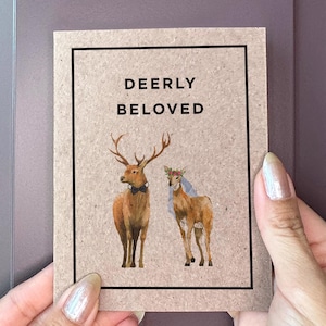 May include: A rectangular card with the words "DEERLY BELOVED" and illustrations of two deer. The deer are brown with antlers and a floral crown. The card is held by a person with manicured nails.