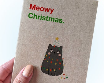 Puede incluir: Una tarjeta marrón con el texto "Meowy Christmas." en rojo y verde. Un gato negro con una estrella en la cabeza está decorado con luces de Navidad.