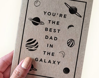 May include: A brown card with black text and illustrations of planets and stars. The text reads "You're the best dad in the galaxy."