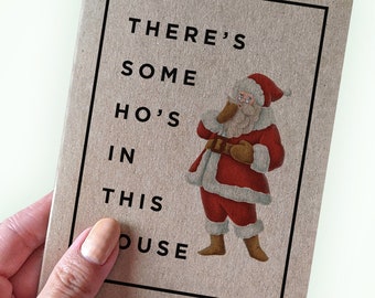 May include: A brown card with a white border and the text "THERE'S SOME HO'S IN THIS OUSE" in black. A cartoon illustration of Santa Claus in a red suit with a white beard and glasses is in the center of the card.