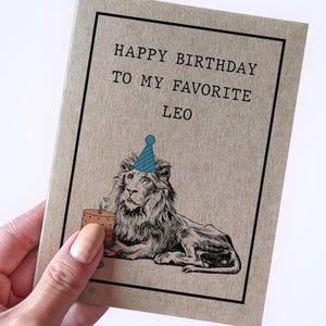 May include: A rectangular greeting card made of light brown paper with a black border. The text reads HAPPY BIRTHDAY TO MY FAVORITE LEO. Below the text is a black and white illustration of a lion wearing a blue party hat next to a small cake.