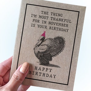 May include: A brown card with a black border and the text "The thing I'm most thankful for in November is your birthday." A black and white illustration of a turkey wearing a pink party hat is below the text. The text "Happy Birthday" is at the bottom of the card.