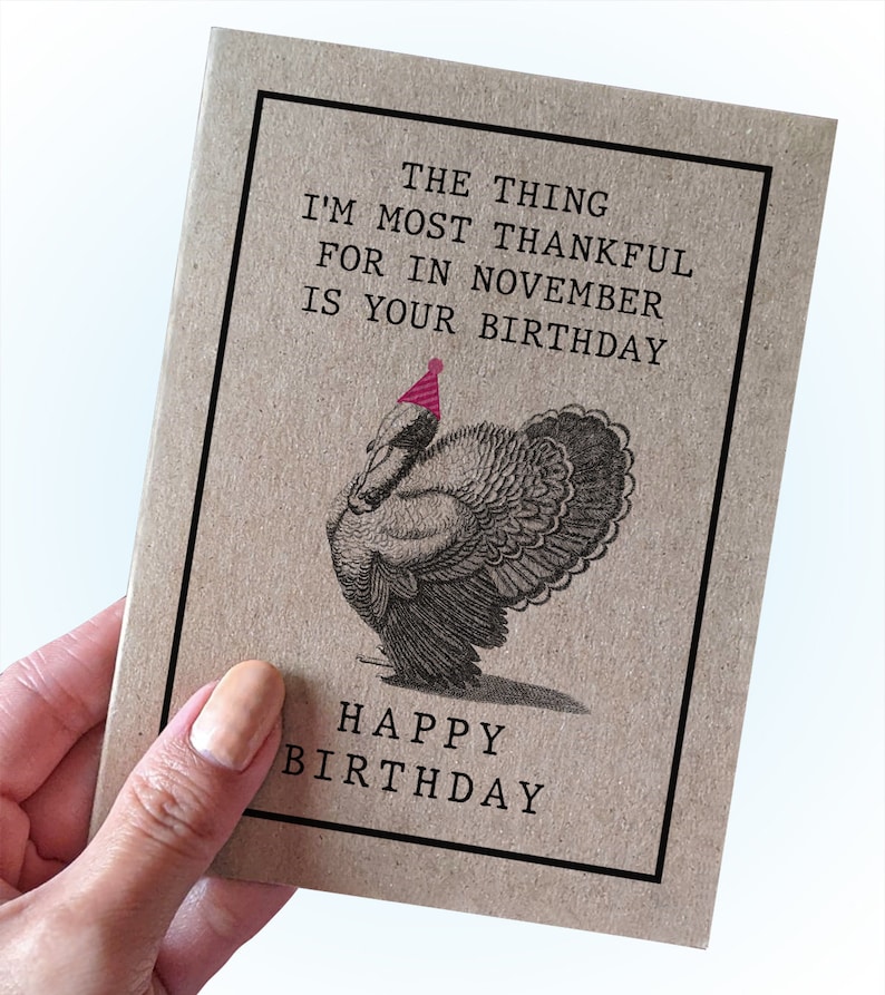 May include: A brown card with a black border and the text "The thing I'm most thankful for in November is your birthday." A black and white illustration of a turkey wearing a pink party hat is below the text. The text "Happy Birthday" is at the bottom of the card.