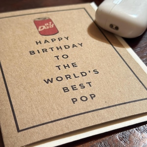 Op de afbeelding: Een bruine kaart met een zwarte rand en de tekst "Happy Birthday to the World's Best Pop" met een rode en witte illustratie van een blikje frisdrank met het woord "Dad" erop.