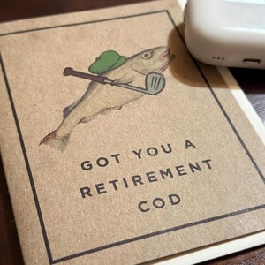 Pu&ograve; includere: Un biglietto marrone con un'illustrazione di un pesce che indossa un cappello da golf verde e tiene un bastone da golf. Il testo sul biglietto dice "Got you a retirement cod."