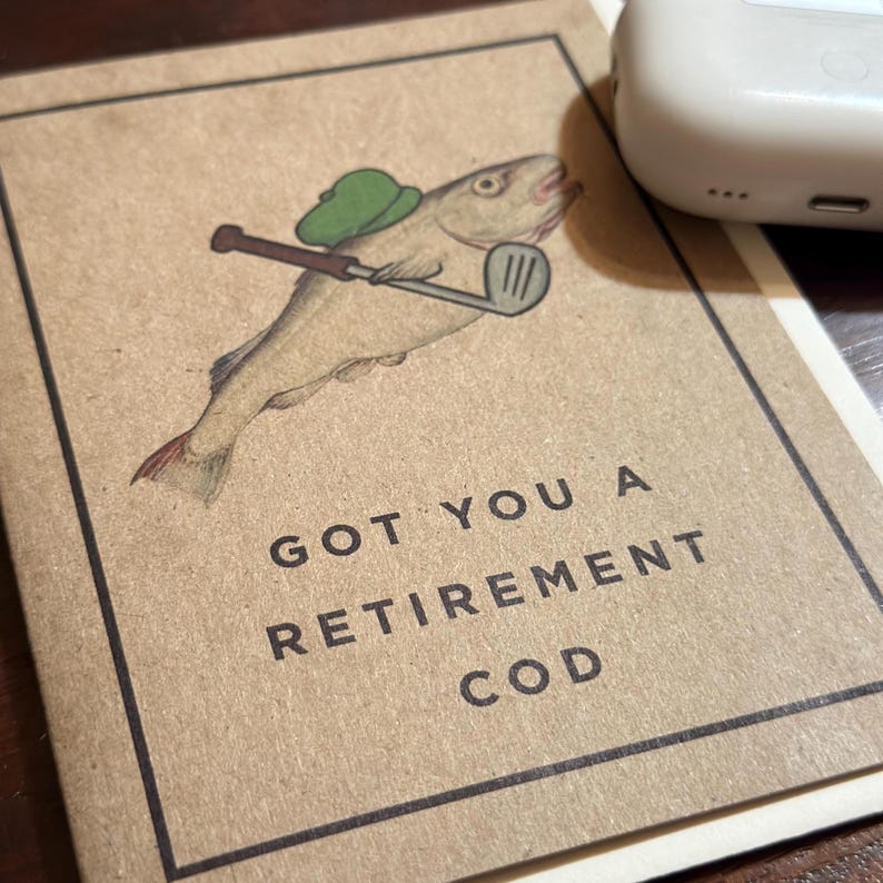 Pu&ograve; includere: Un biglietto marrone con un'illustrazione di un pesce che indossa un cappello da golf verde e tiene un bastone da golf. Il testo sul biglietto dice "Got you a retirement cod."