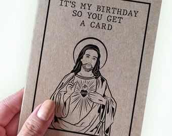 Pode incluir: Um cartão de aniversário castanho com texto preto que diz "IT'S MY BIRTHDAY SO YOU GET A CARD." O cartão apresenta uma ilustração a preto e branco de Jesus com um símbolo de coração. O cartão é segurado por uma pessoa.