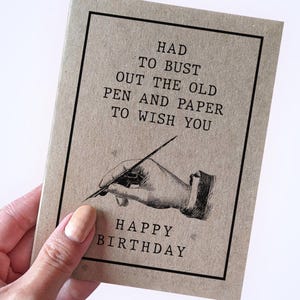May include: A rectangular greeting card made of light brown kraft paper with a black border. The text reads: HAD TO BUST OUT THE OLD PEN AND PAPER TO WISH YOU HAPPY BIRTHDAY. Below the text is a black and white illustration of a hand holding a pen.