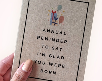 May include: A brown card with a black border and a birthday-themed illustration of presents and balloons. The text on the card reads "ANNUAL REMINDER TO SAY I'M GLAD YOU WERE BORN."
