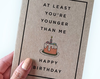 May include: A brown card with black text that reads "At least you're younger than me."  A cartoon cake with a single candle is below the text. The text "Happy Birthday" is below the cake.