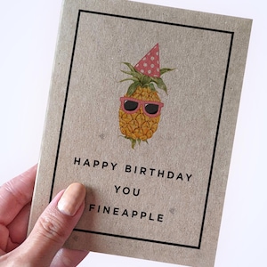 May include: A tan greeting card featuring a central illustration of a yellow pineapple wearing a pink polka-dot party hat and pink sunglasses. Below the pineapple, the text reads HAPPY BIRTHDAY YOU FINEAPPLE in black, all enclosed in a black border.