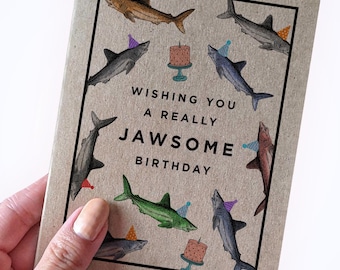 May include: A brown card with a birthday greeting that says "Wishing you a really Jawsome Birthday". The card is decorated with colorful sharks wearing party hats and a cake.