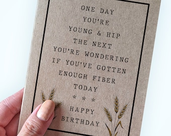 May include: A brown card with a humorous birthday message that reads "One day you're young & hip the next you're wondering if you've gotten enough fibre today*** Happy Birthday". The card has a brown background with a black border and a small illustration of wheat stalks in the bottom right corner.