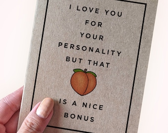 Pode incluir: Um cartão com fundo castanho e borda preta. O texto diz "I LOVE YOU FOR YOUR PERSONALITY BUT THAT IS A NICE BONUS" com uma ilustração de pêssego. O cartão é segurado por uma pessoa.