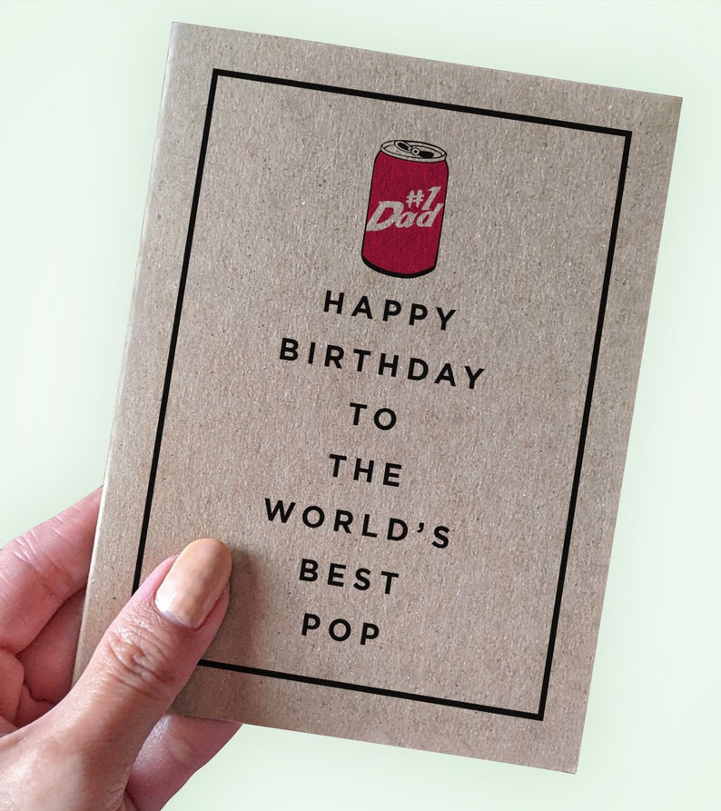 Op de afbeelding: Een bruine kaart met een zwarte rand en de tekst "Happy Birthday to the World's Best Pop" in zwart. Bovenaan de kaart staat een rode blik met de tekst "#1 Dad".