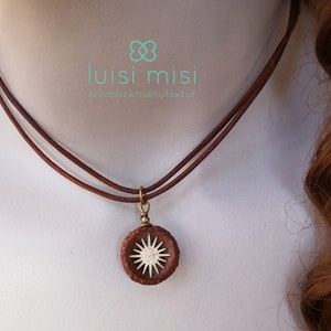 Op de afbeelding: Een bruin leren ketting met een hanger met een sunburst-ontwerp. De hanger is cirkelvormig met een lichtgekleurd sterrenpatroon in een donkerbruin frame. De ketting heeft twee dunne bruine koorden. De merknaam "luisi misi" is zichtbaar.