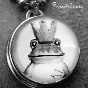Könnte beinhalten: Eine silberne Kettenkette mit einem runden Anhänger, der ein Schwarzweißbild eines Frosches mit einer Krone zeigt. Der Anhänger trägt die Aufschrift "Froschkönig" und "luisi misi unikatschmuck mit" in schwarzer Schrift.