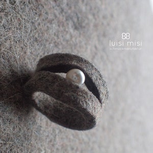 May include: A grey felt ring with a white pearl nestled inside. The ring is made by luisi misi schmuckmanufaktur.