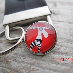 May include: A silver key chain with a red and white moose design on a round glass charm. The charm says "luisi misi unikatschmuck".