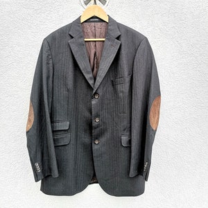 Puede incluir: Una chaqueta gris oscura con un patrón de espiga y parches de cuero marrón en los codos. La chaqueta tiene un cierre de tres botones y está colgada de una percha de madera.