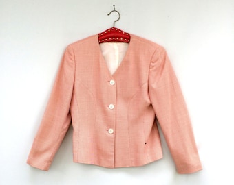 Puede incluir: Una chaqueta de color rosa pálido con cuello en V y cuatro botones blancos. La chaqueta está colgada en una percha roja con tachuelas plateadas.