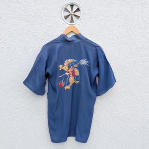 May include: A blue kimono-style robe with a dragon embroidery on the back. The dragon is depicted in gold, red, and blue, with intricate details. The robe has short sleeves and hangs on a wooden hanger against a white wall.