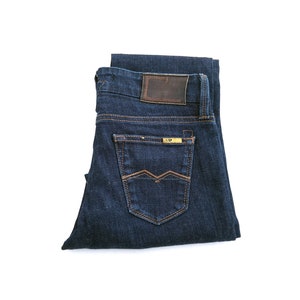May include: A pair of dark blue jeans folded in half, showing the back pockets. The jeans have a brown leather patch with stitching on the back waistband and a gold label on the right back pocket.